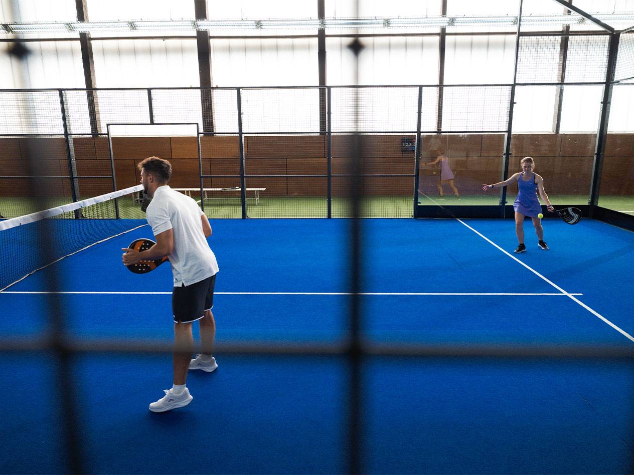 Due persone giocano a padel su un campo blu, l'uomo tiene una racchetta.