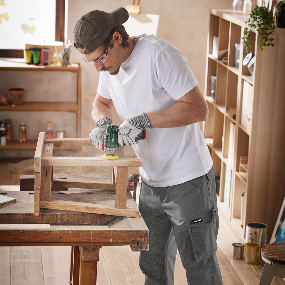 Homme ponçant des meubles en bois avec un outil électrique, portant un pantalon de travail et un équipement de protection.