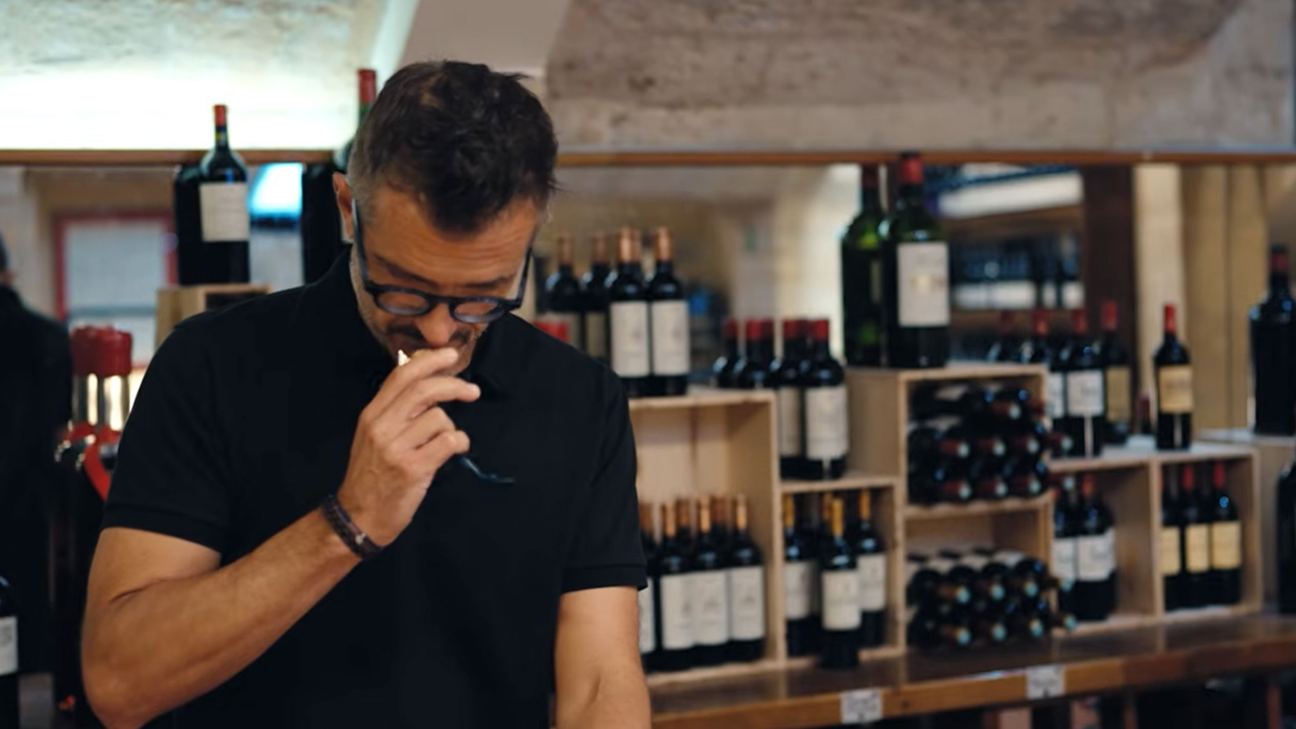 Homme dans une cave à vin dégustant du vin, entouré de bouteilles sur des étagères en bois.
