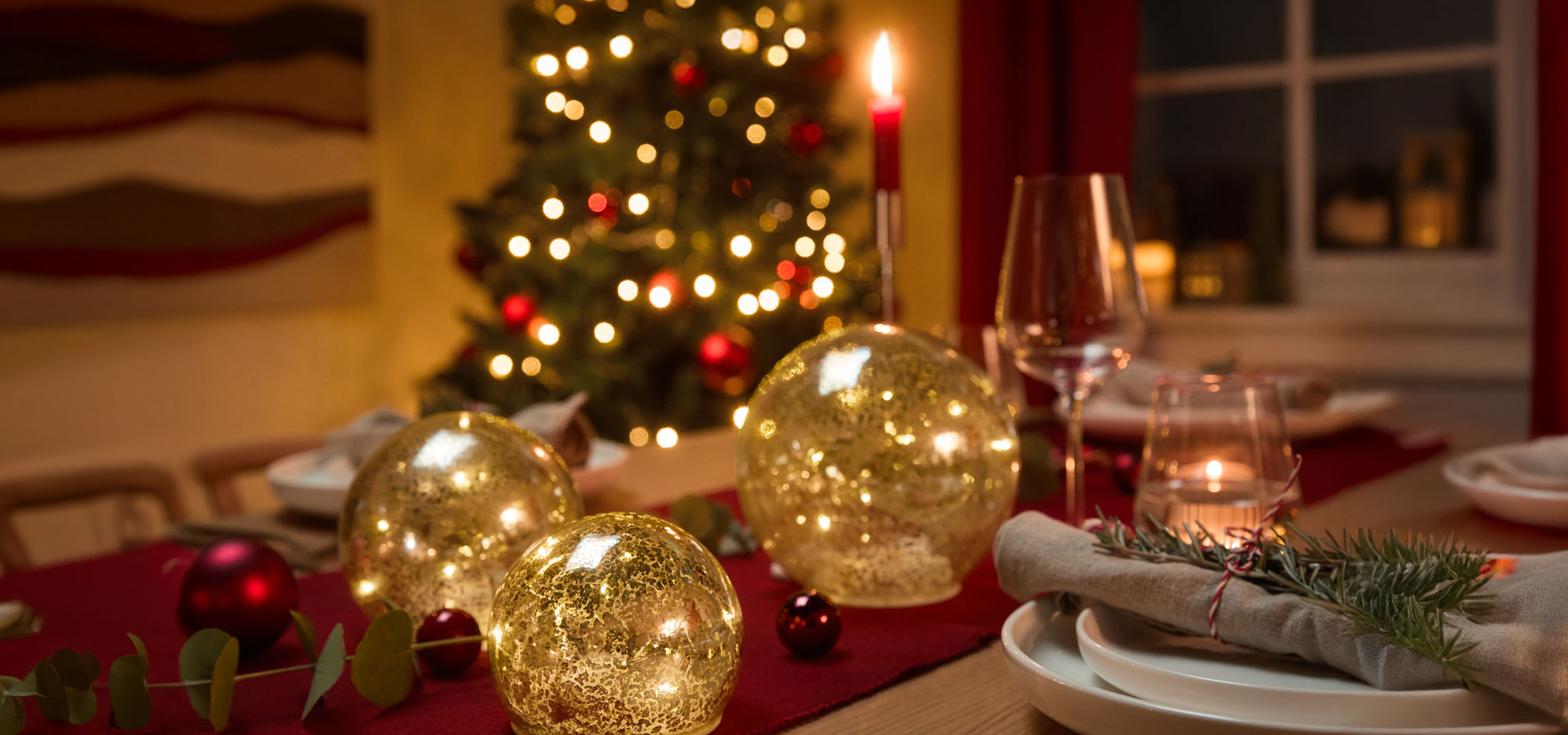Weihnachtlich gedeckter Tisch mit goldenen Leuchtkugeln, roten Kugeln und Tannenzweigen.
