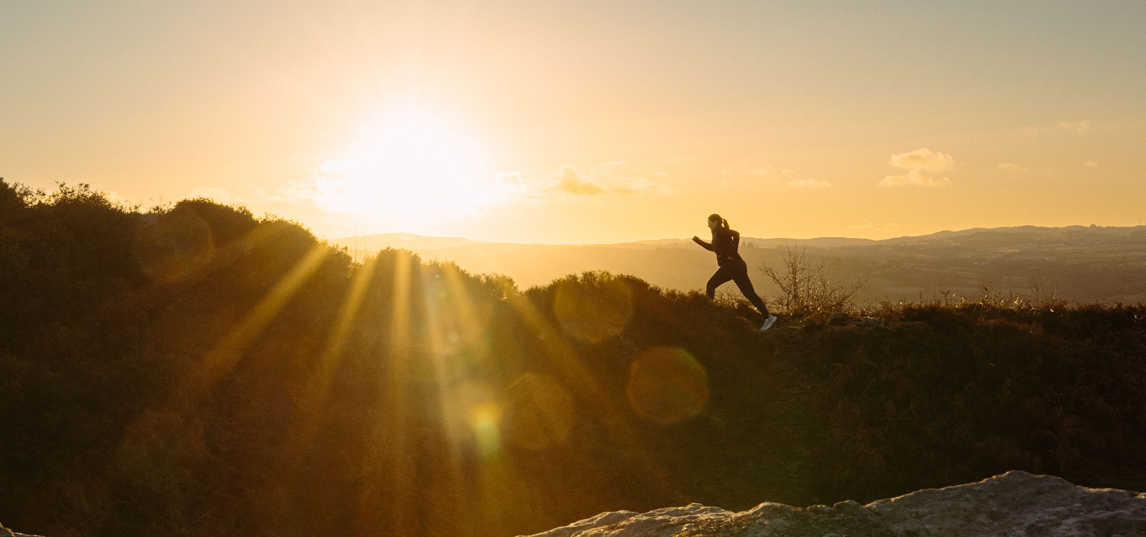 Frau läuft auf einem Hügel bei Sonnenuntergang, mit Sonnenstrahlen und einer malerischen Landschaft.