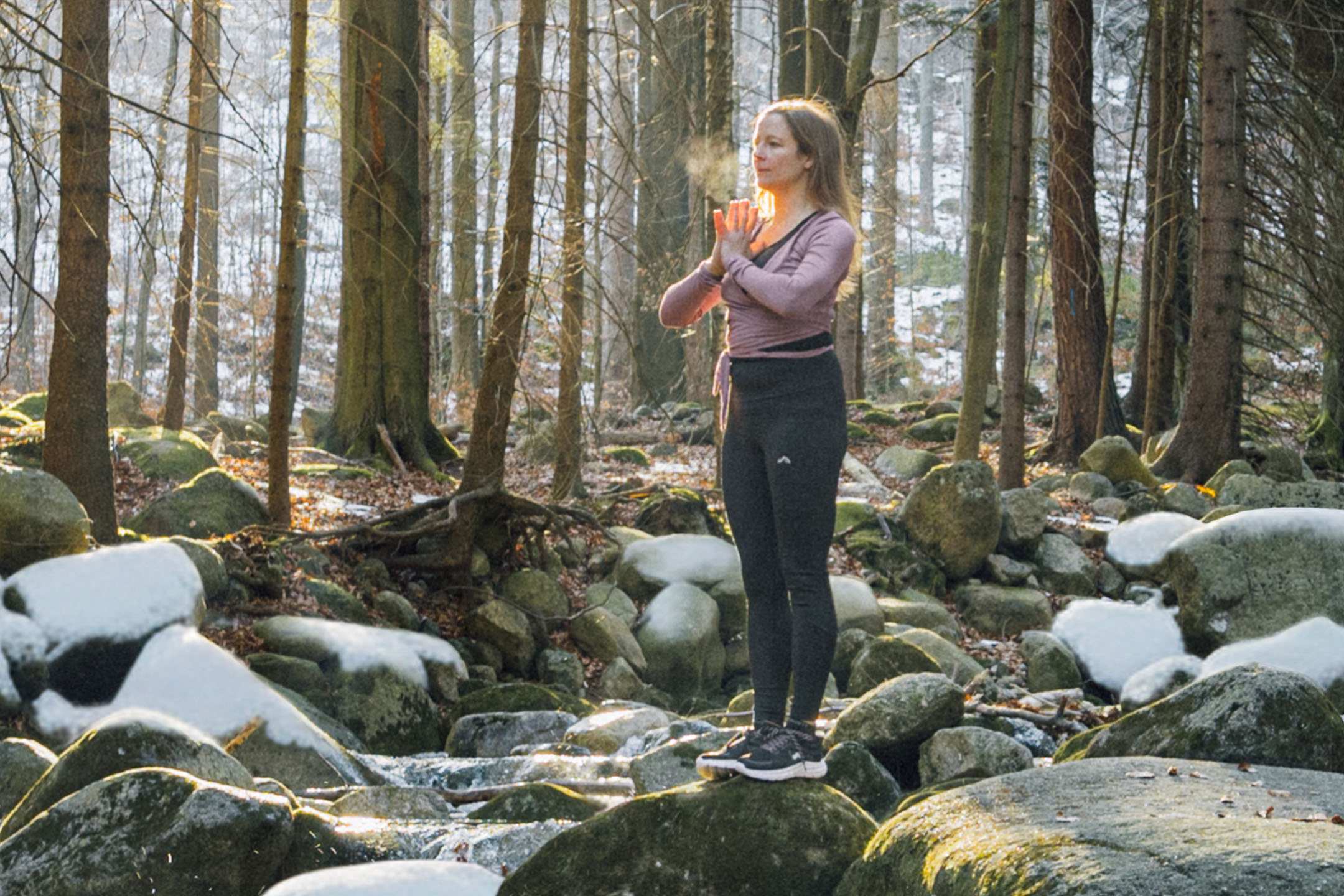 Frau in Sportkleidung macht Yoga in einem Wald mit schneebedeckten Felsen und Bäumen.
