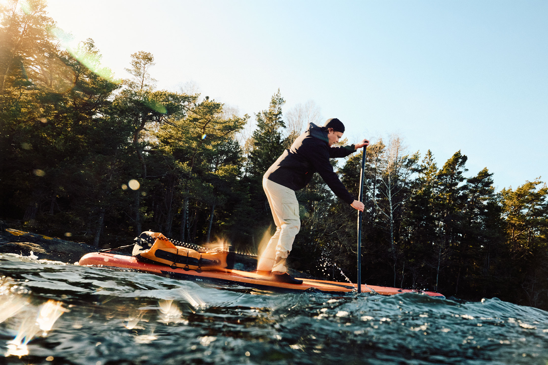 Mann in beiger Hose und schwarzer Jacke paddelt auf einem orangefarbenen Paddleboard