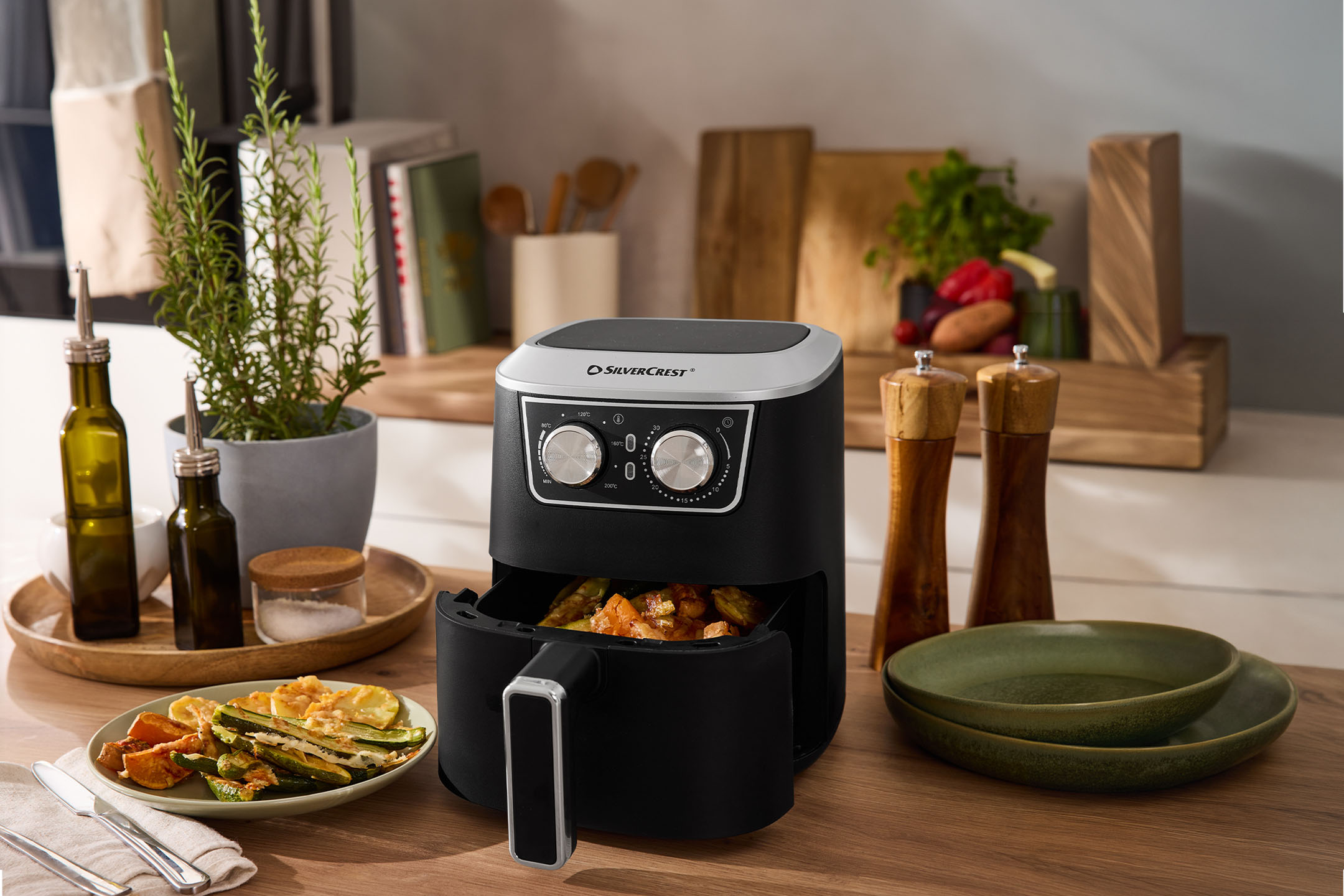 Friteuse à air noire avec légumes, épices et vaisselle sur un comptoir en bois.