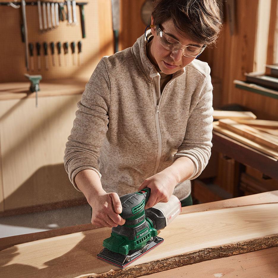 Frau mit Elektroschleifer schleift ein Holzstück in einer Werkstatt.