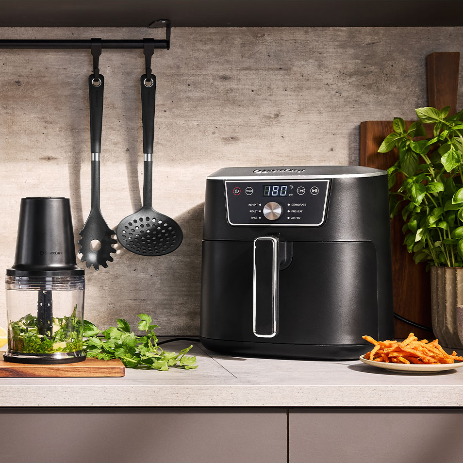 SilverCrest Heißluftfritteuse und Zerkleinerer auf einer Küchenzeile mit frischen Kräutern und Süßkartoffelpommes.