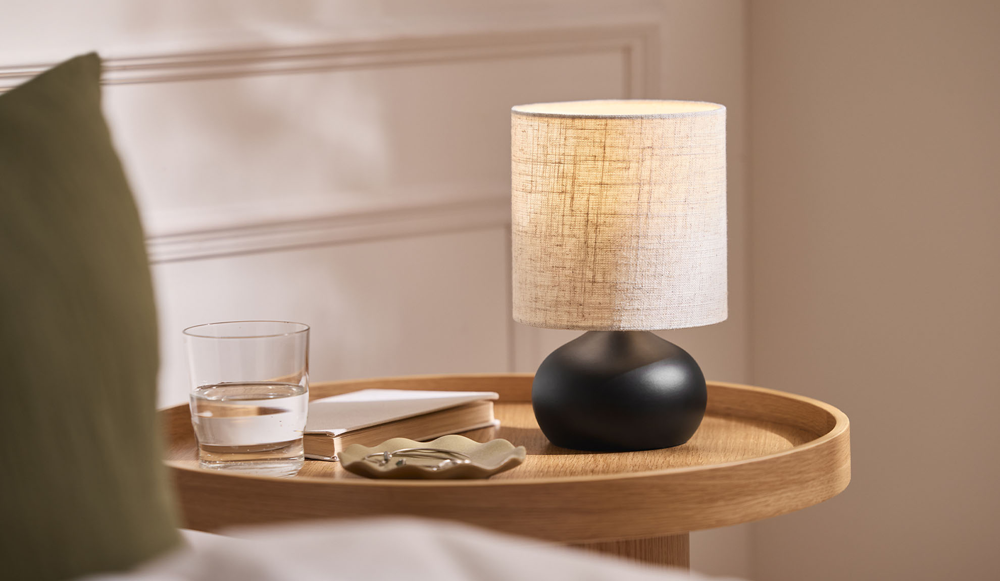 Lampada da comodino, bicchiere, libro e vassoio portagioie su un vassoio di legno accanto al letto.