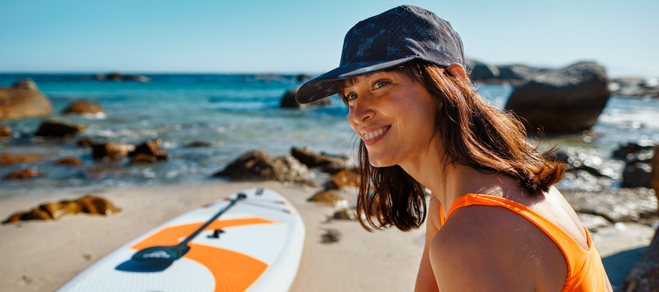 Femme souriante en maillot de bain orange et casquette de baseball, près d'une planche de paddle surf sur la plage.