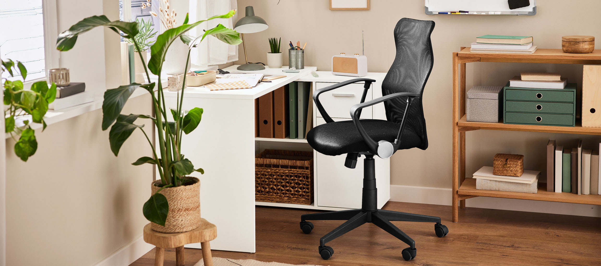 Ein Heimbüro mit einem weißen Schreibtisch, einem schwarzen Bürostuhl und einem Holzregal.