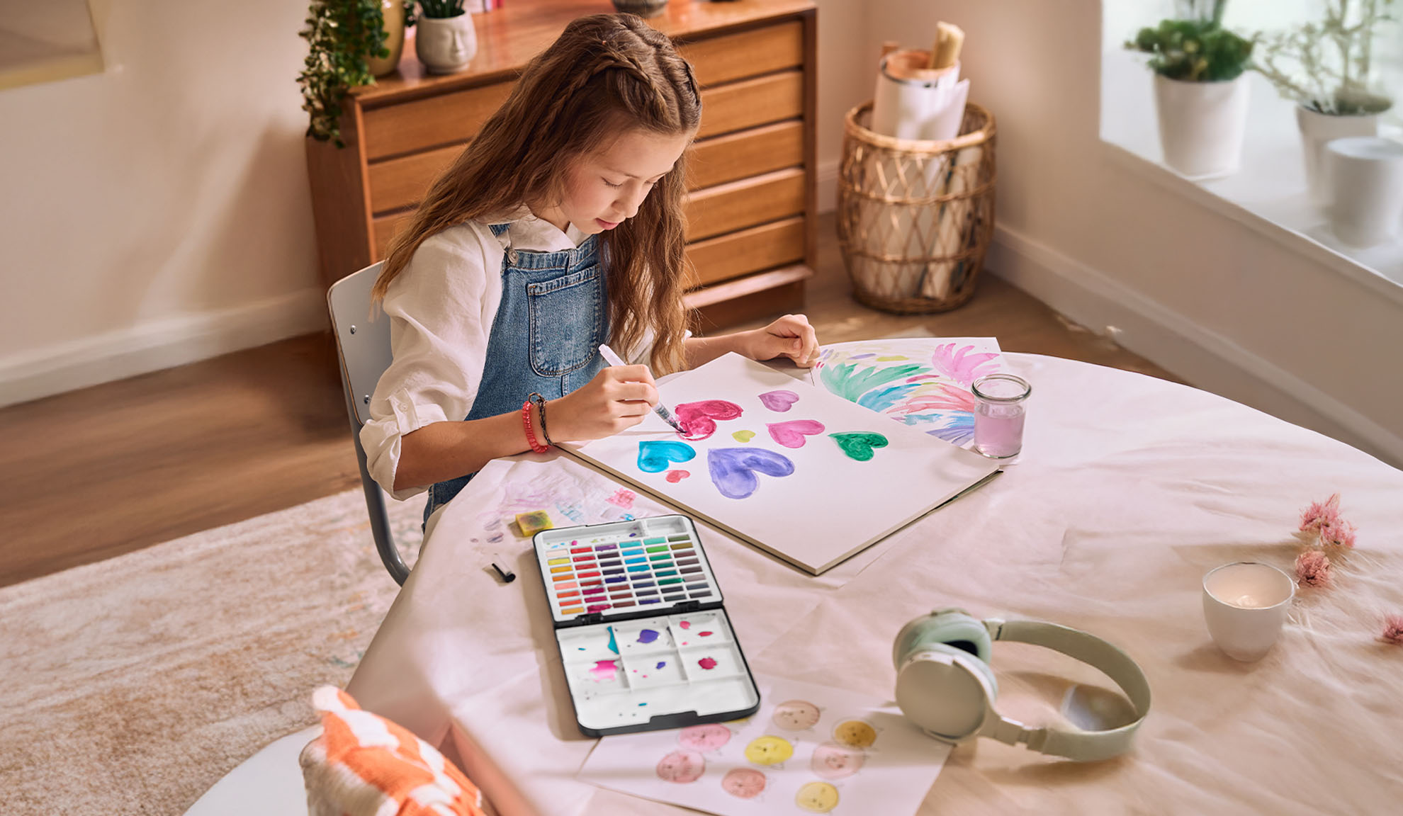 Une fille peignant des cœurs à l'aquarelle à une table, avec un set d'aquarelle et des écouteurs à proximité.