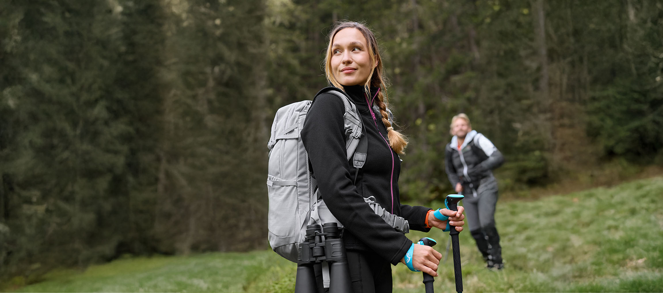 Frau mit Rucksack und Wanderstöcken, mit einem Mann im Hintergrund, im Wald.