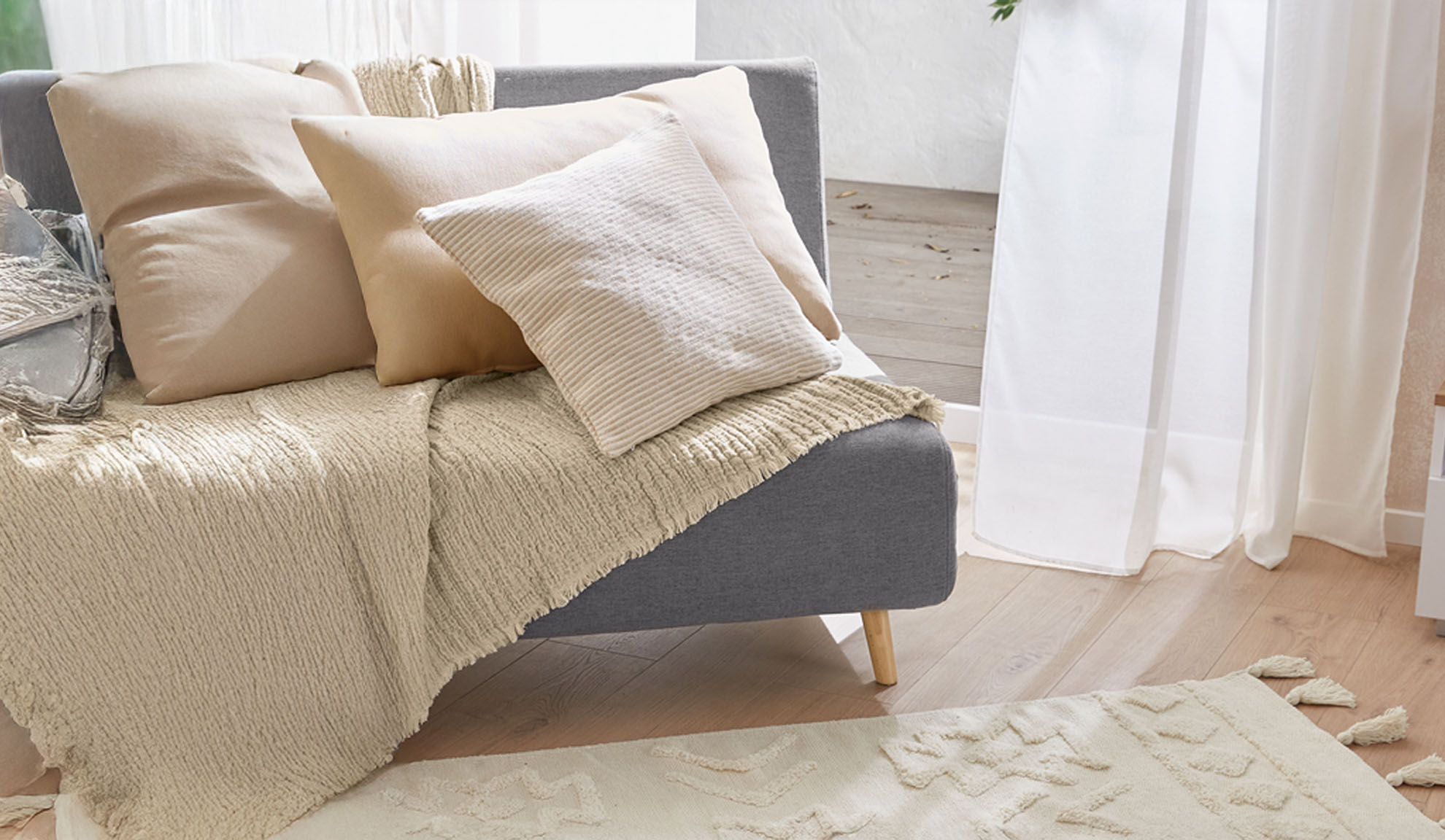 Graues Sofa mit beigen Kissen und strukturierter Decke in einem hellen Wohnzimmer.
