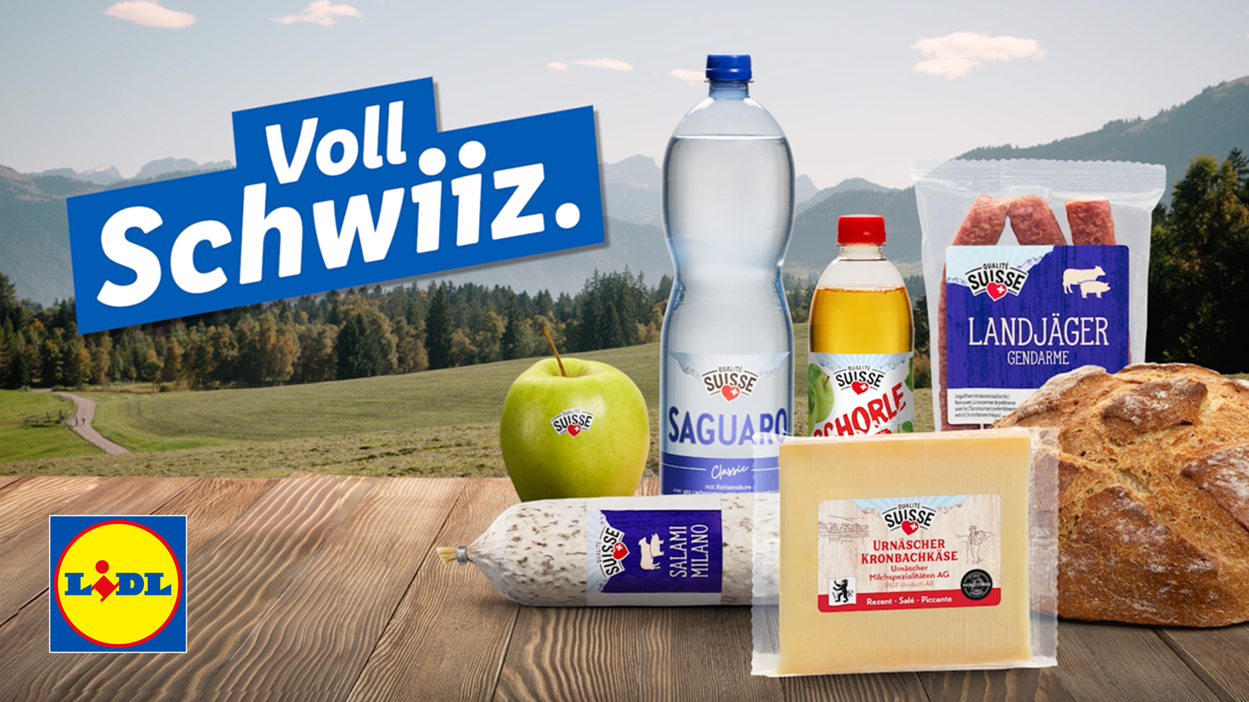 Schweizer Produkte auf einem Holztisch vor Bergkulisse, darunter Käse, Salami, Brot, Wasser und Apfelschorle.