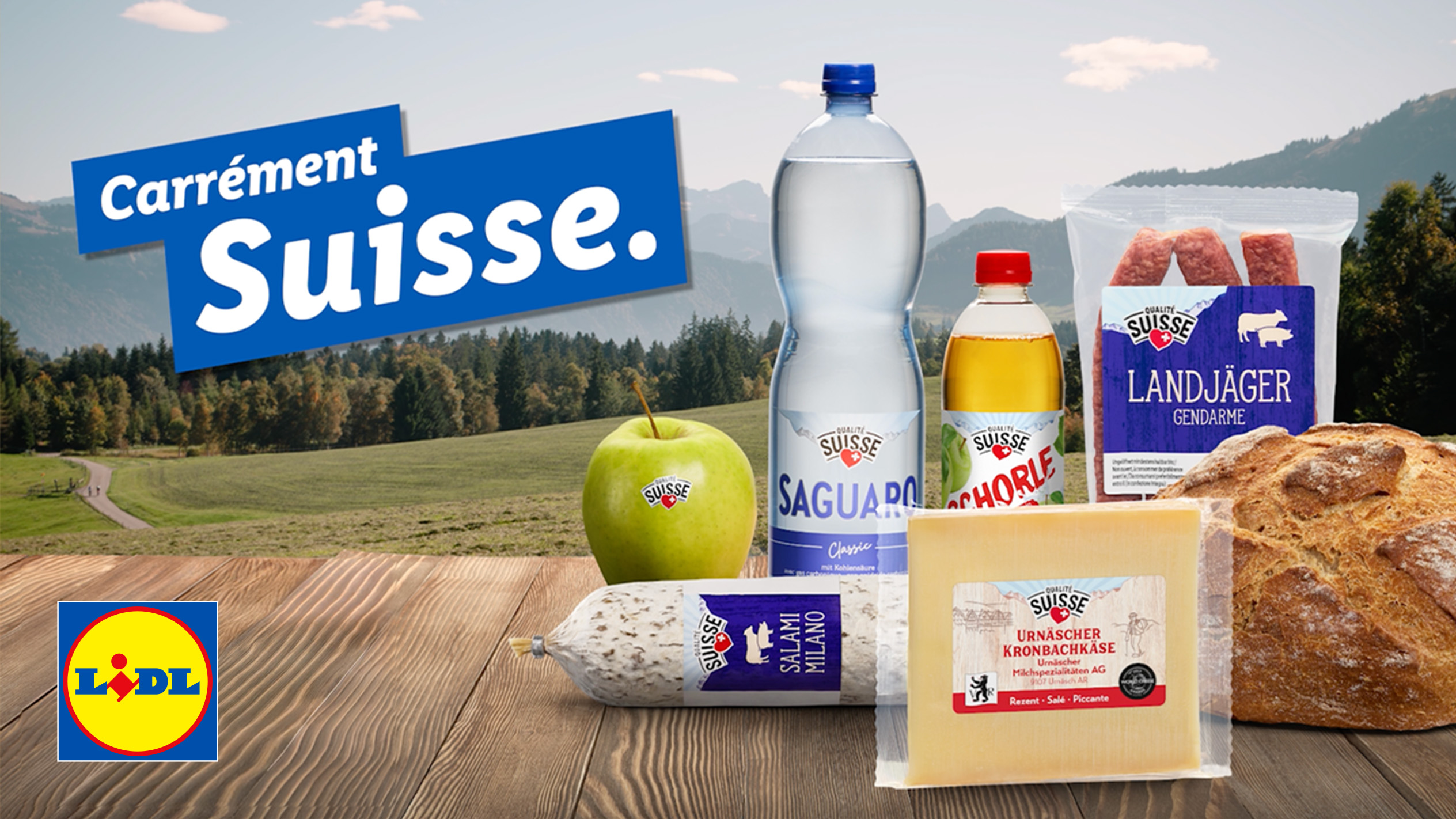 Produits suisses sur une table en bois avec un fond de montagne : saucisson, fromage, eau, jus de pomme, pomme, pain et saucisses.