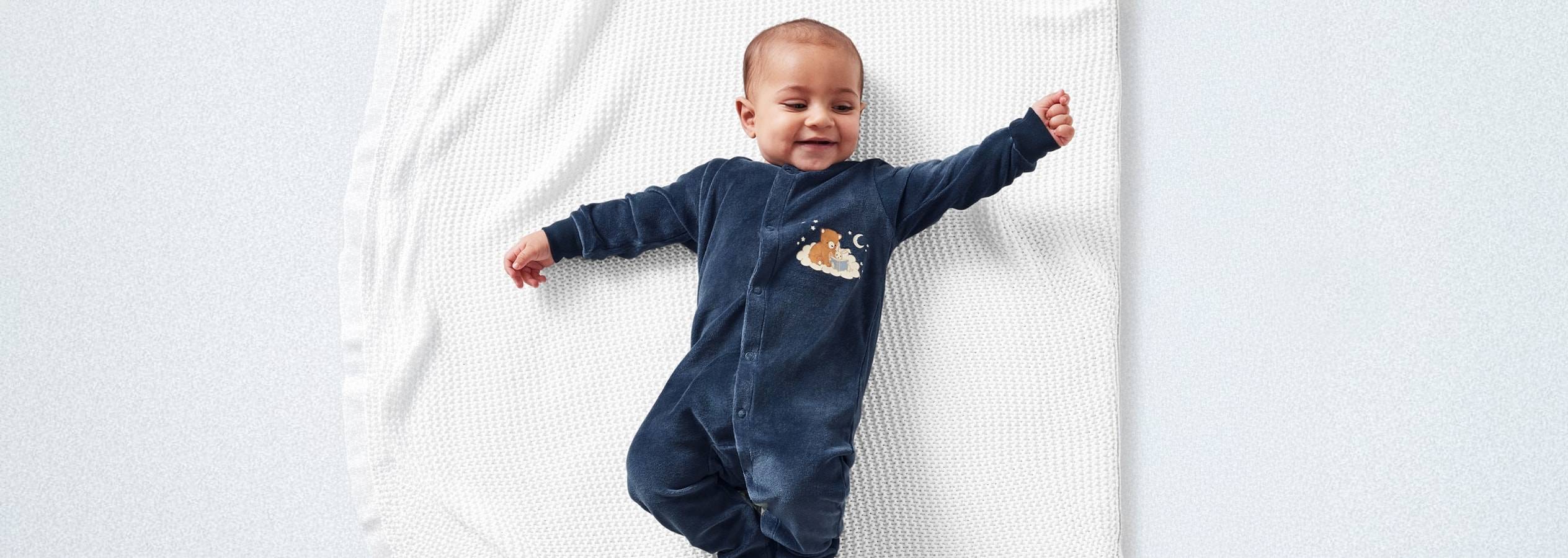 Un bambino sorridente sdraiato su una coperta bianca, indossa un pigiamino blu scuro con un orsetto.