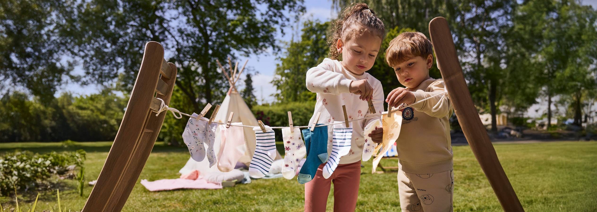 Zwei Kinder hängen bunte Socken an einer Spielzeug-Wäscheleine im Garten auf.