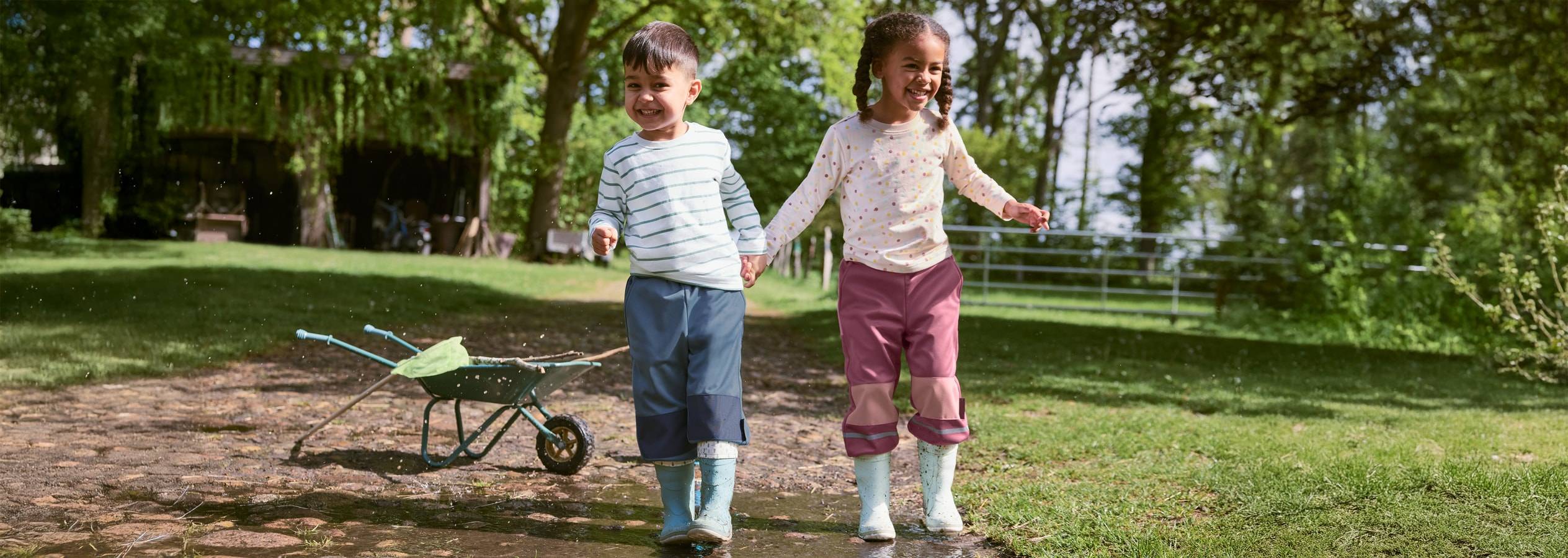 Deux enfants jouent avec une brouette à la campagne, portant des bottes et des pantalons imperméables.
