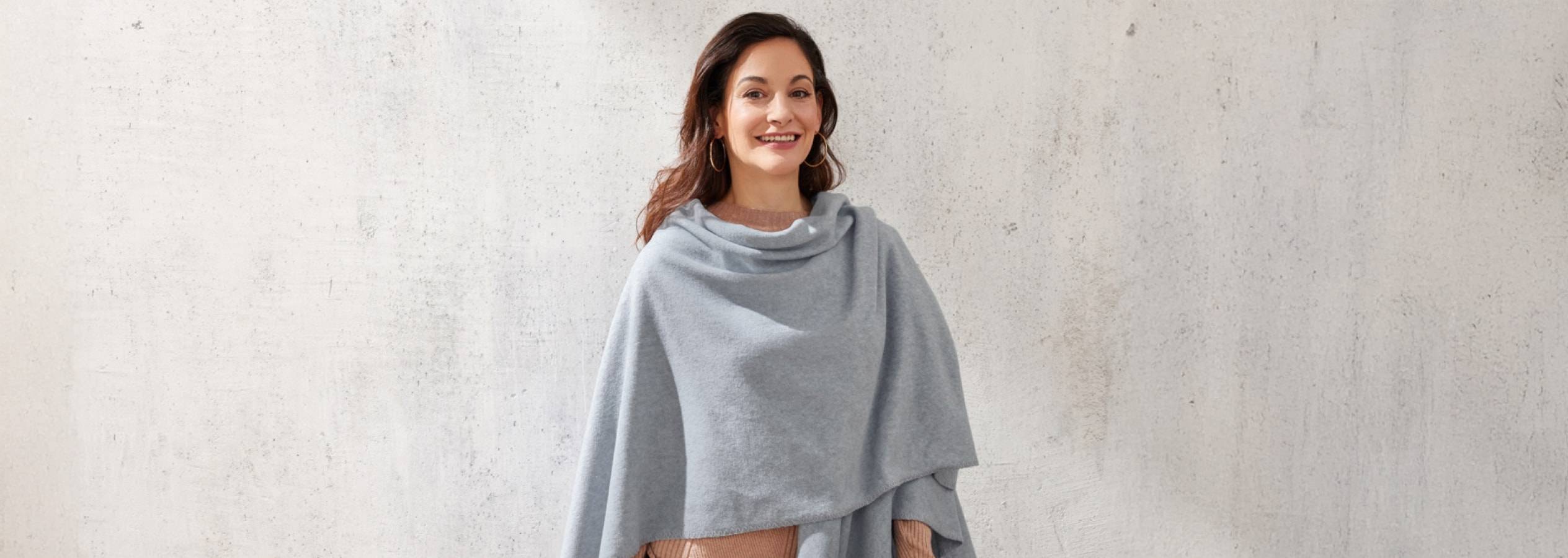 Eine lächelnde Frau trägt einen grauen Poncho und goldene Creolen.