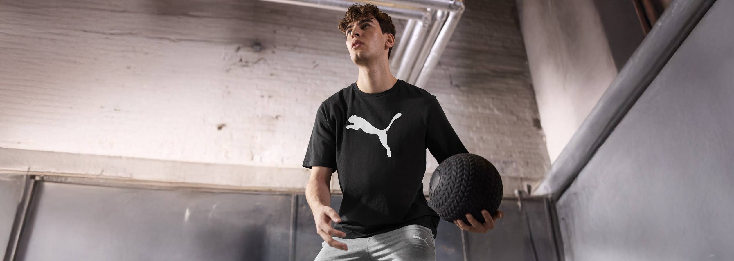 Ein Mann trägt ein schwarzes Puma-T-Shirt und graue Hosen und hält einen Medizinball.
