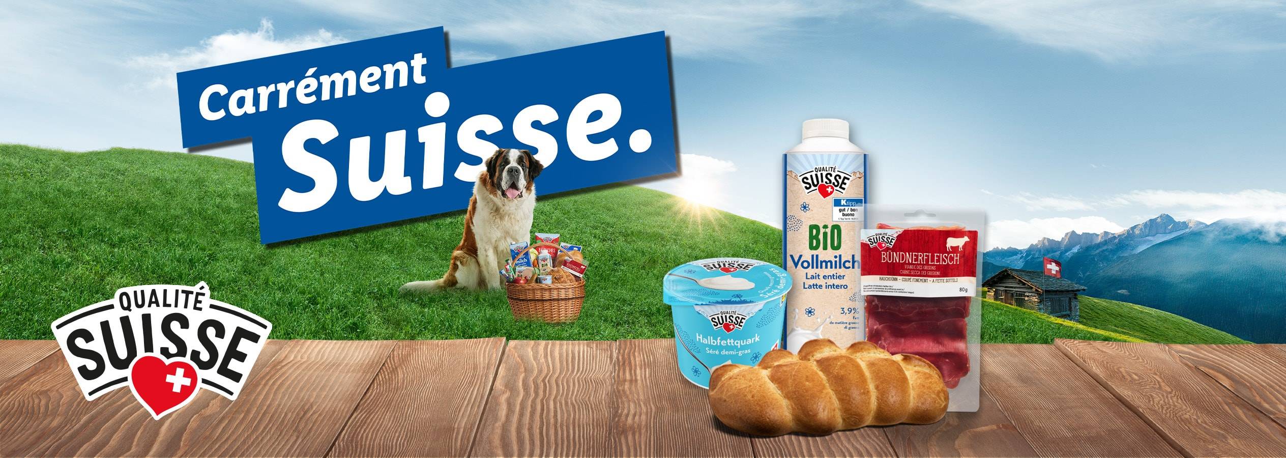 Produits suisses comme du lait, du séré, de la viande des Grisons et du pain sur une table en bois devant un paysage de montagne avec un Saint-Bernard.