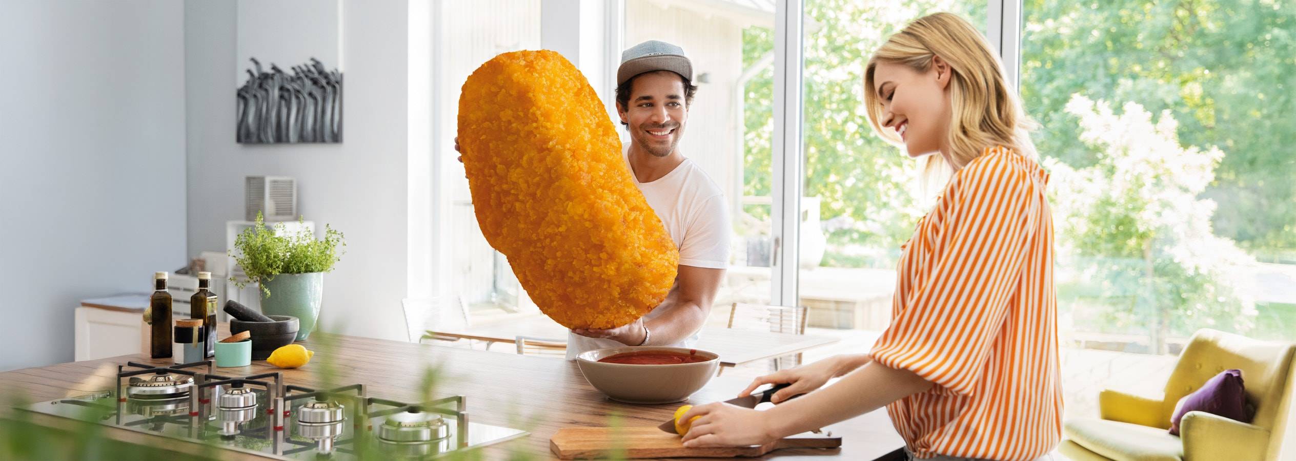 Ein Mann hält eine riesige Hähnchenkrokette, während eine Frau in einer hellen Küche eine Zitrone schneidet.