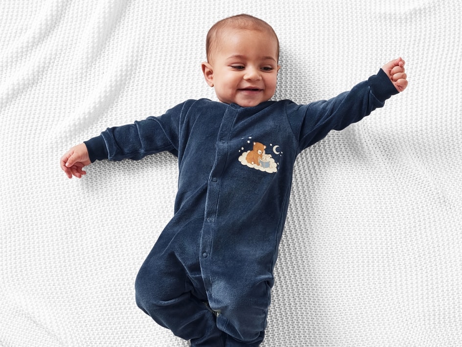 Lächelndes Baby auf einer weißen Decke, trägt einen dunkelblauen Schlafanzug mit Bärenmotiv