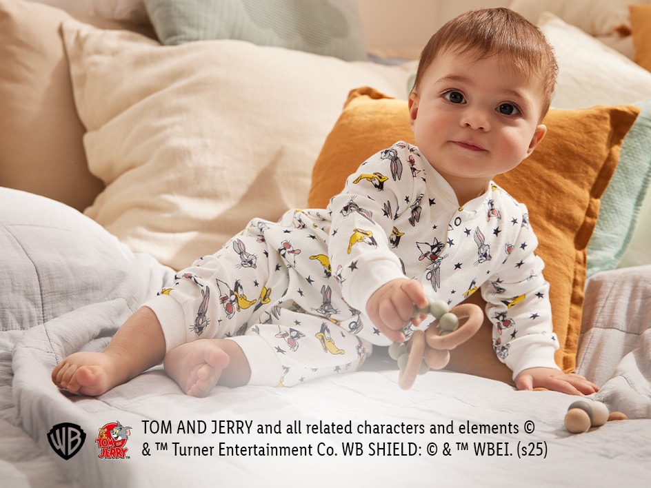 Ein Baby in einem Tom und Jerry Strampler spielt mit einem Beißring auf einem Bett.