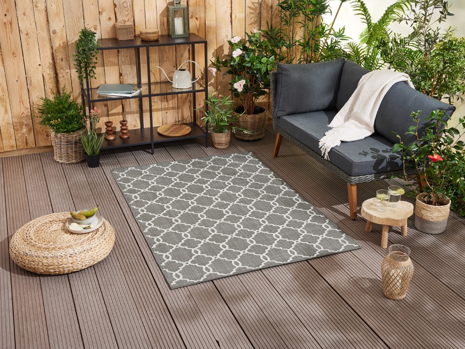 Ein hellgrauer Teppich mit geometrischem Muster schmückt eine Terrasse mit Sofa, Pflanzen und Deko-Accessoires.