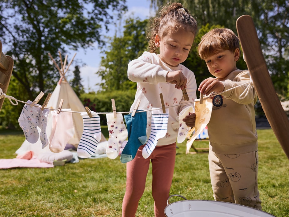 Zwei Kinder hängen Socken und Stoffe an einer Spielzeug-Wäscheleine im Freien auf.