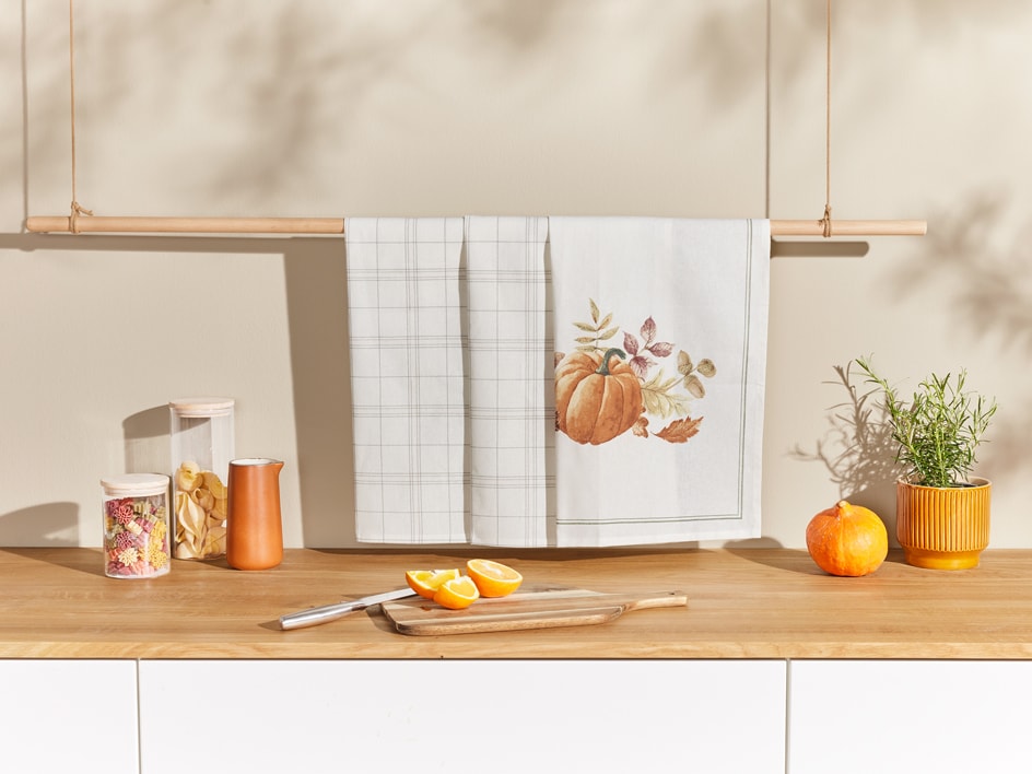 Due strofinacci da cucina appesi a un bastone di legno, con decorazioni autunnali e alimenti.