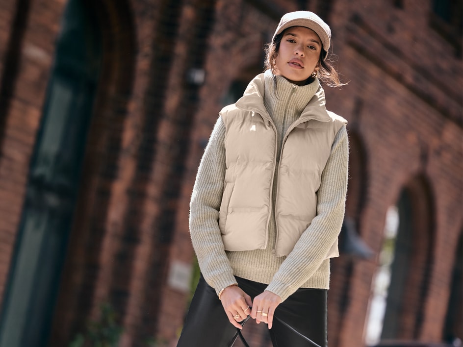 Donna in gilet imbottito beige, maglione a collo alto e pantaloni neri.