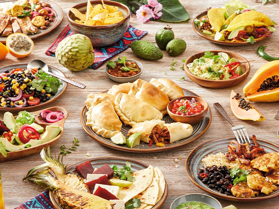Ein Holztisch mit einer Vielzahl lateinamerikanischer Gerichte, darunter Empanadas, Tacos, Salate und Obst.