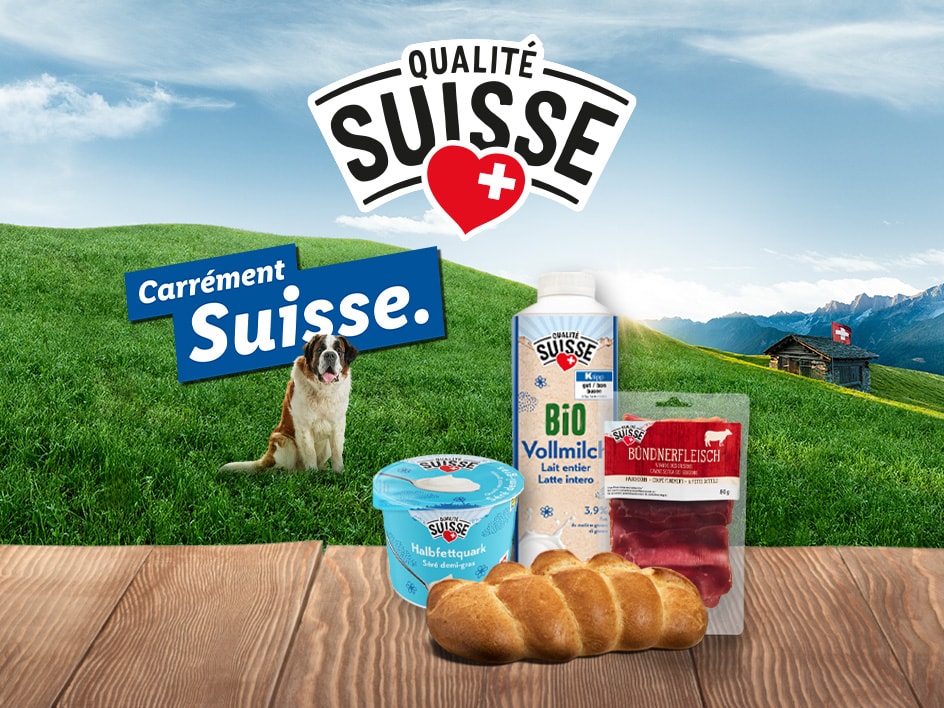 Produits Qualité Suisse comme du lait, de la viande des Grisons et du séré sur une table en bois devant un paysage de montagne suisse avec un Saint-Bernard.