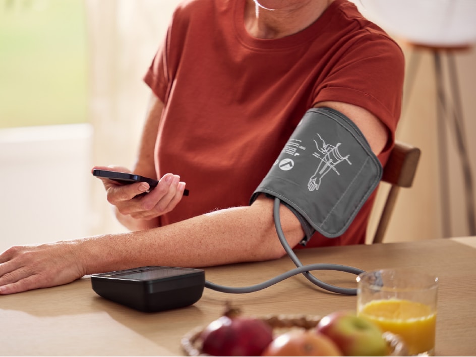 Eine Person misst ihren Blutdruck mit einem Oberarm-Blutdruckmessgerät und hält dabei ein Smartphone.