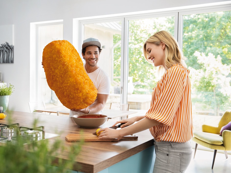 Ein Mann hält eine riesige panierte Hähnchenkrokette, während eine Frau in einer hellen Küche eine Zitrone schneidet.