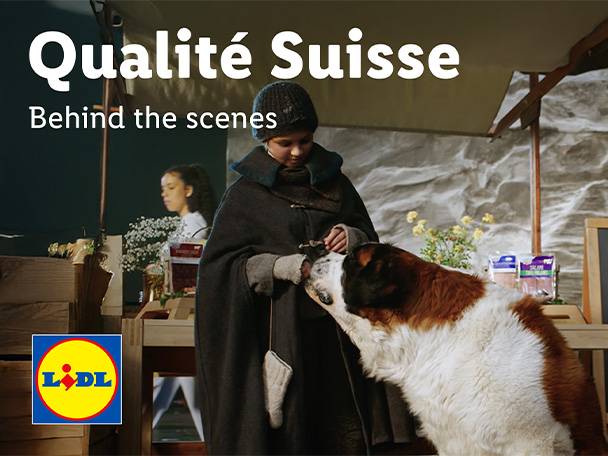 Qualité Suisse: Hinter den Kulissen. Ein Kind in traditioneller Kleidung streichelt einen Bernhardiner, mit verpackten Produkten im Hintergrund.