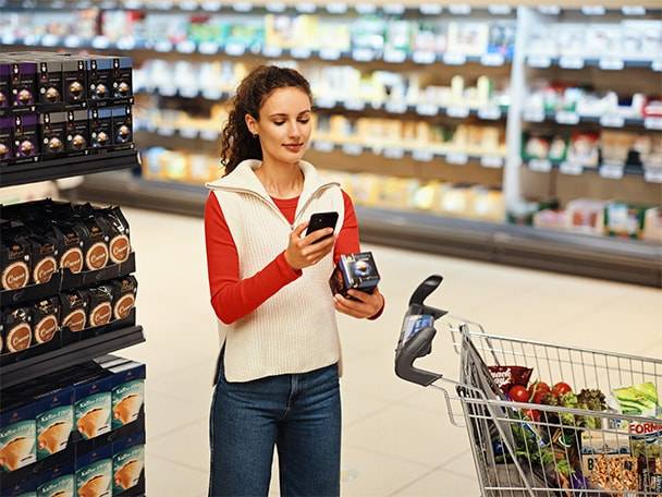 Frau scannt ein Kaffeeprodukt mit ihrem Smartphone, mit einem Einkaufswagen voller Lebensmittel.