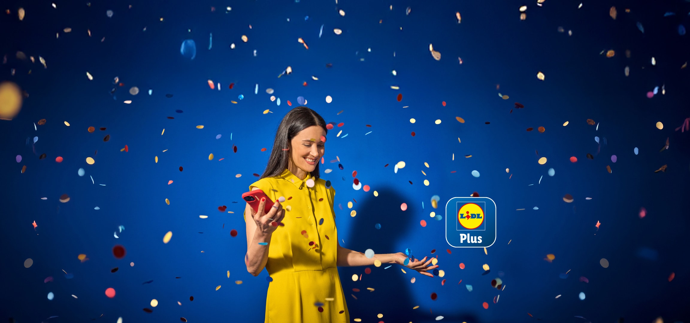 Una donna sorridente in abito giallo, che tiene un telefono rosso, sotto una pioggia di coriandoli colorati.