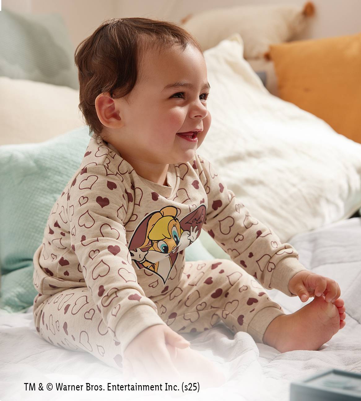 Lächelndes Baby in Pyjama mit Herz- und Lola Bunny-Aufdruck, auf einem Bett sitzend.