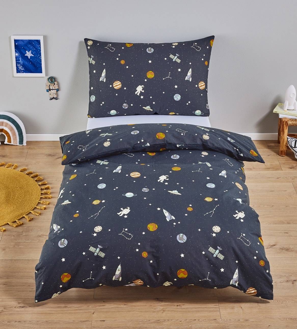 Un lit d'enfant avec une parure de lit sur le thème de l'espace, avec des planètes, des fusées et des astronautes.