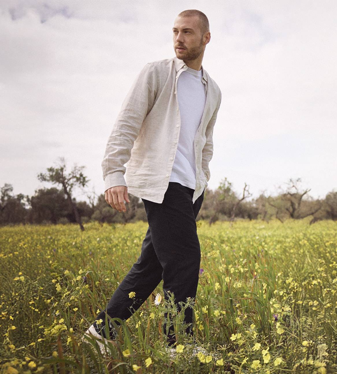 Un homme marche dans un champ fleuri, vêtu d'une chemise en lin beige et d'un pantalon noir.