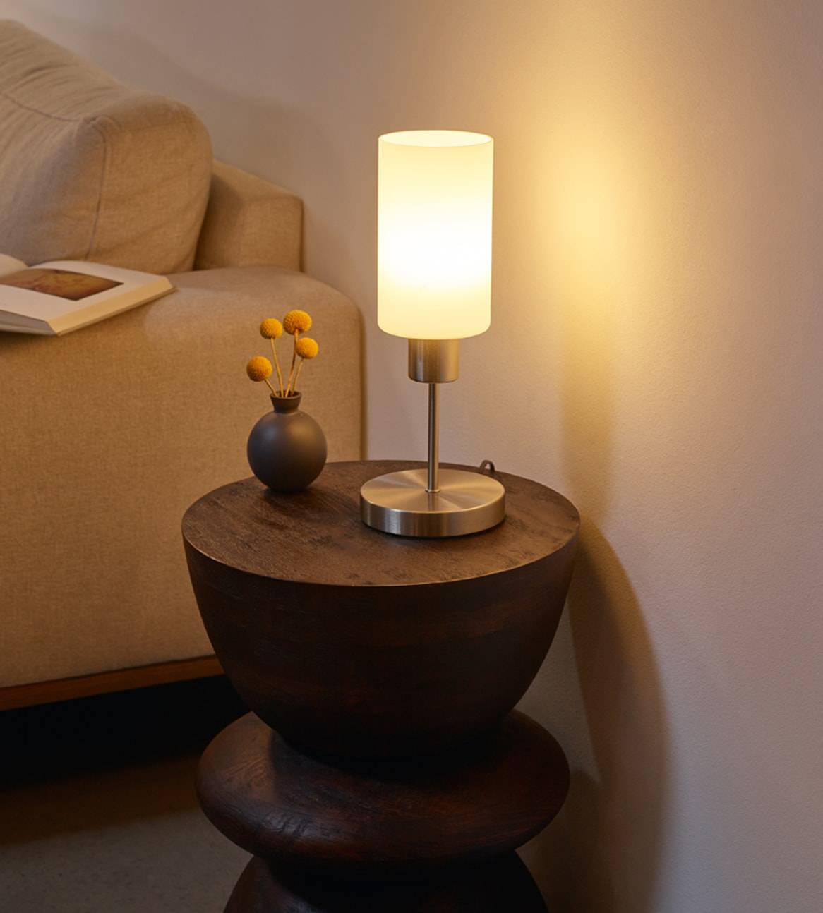 Eine moderne Tischlampe mit opalweißem Glasschirm steht auf einem dunklen Holztischchen.