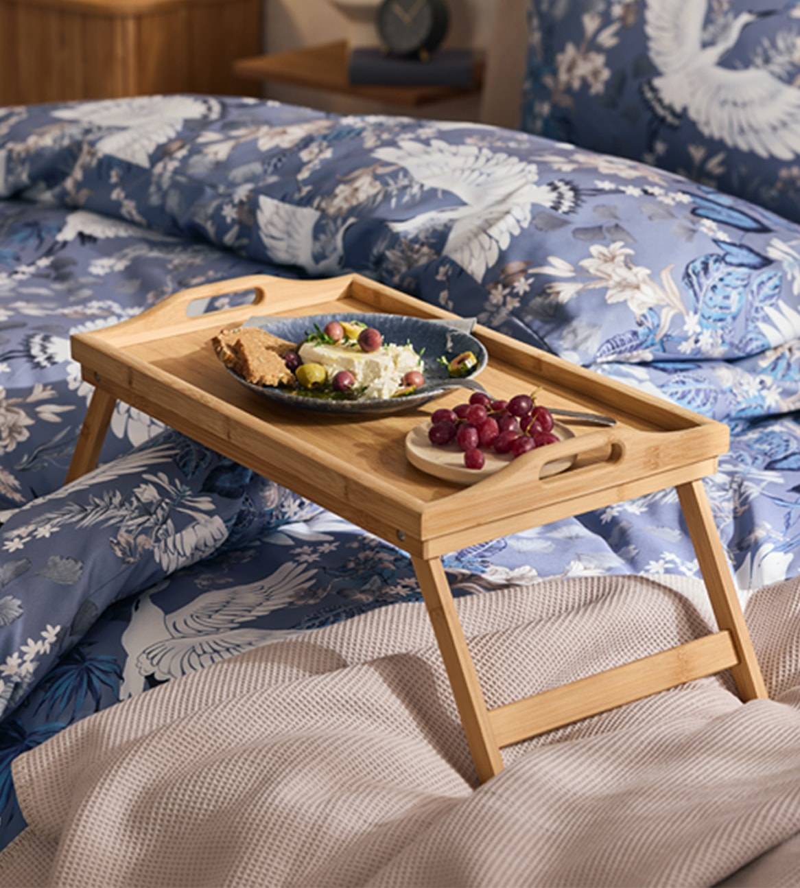 Un vassoio da letto in bambù con colazione su biancheria da letto blu a motivi di uccelli.