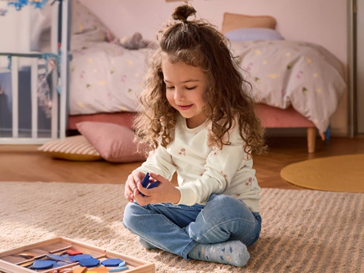 Ein kleines Mädchen spielt mit einem Holzpuzzle auf dem Teppich in einem Schlafzimmer.