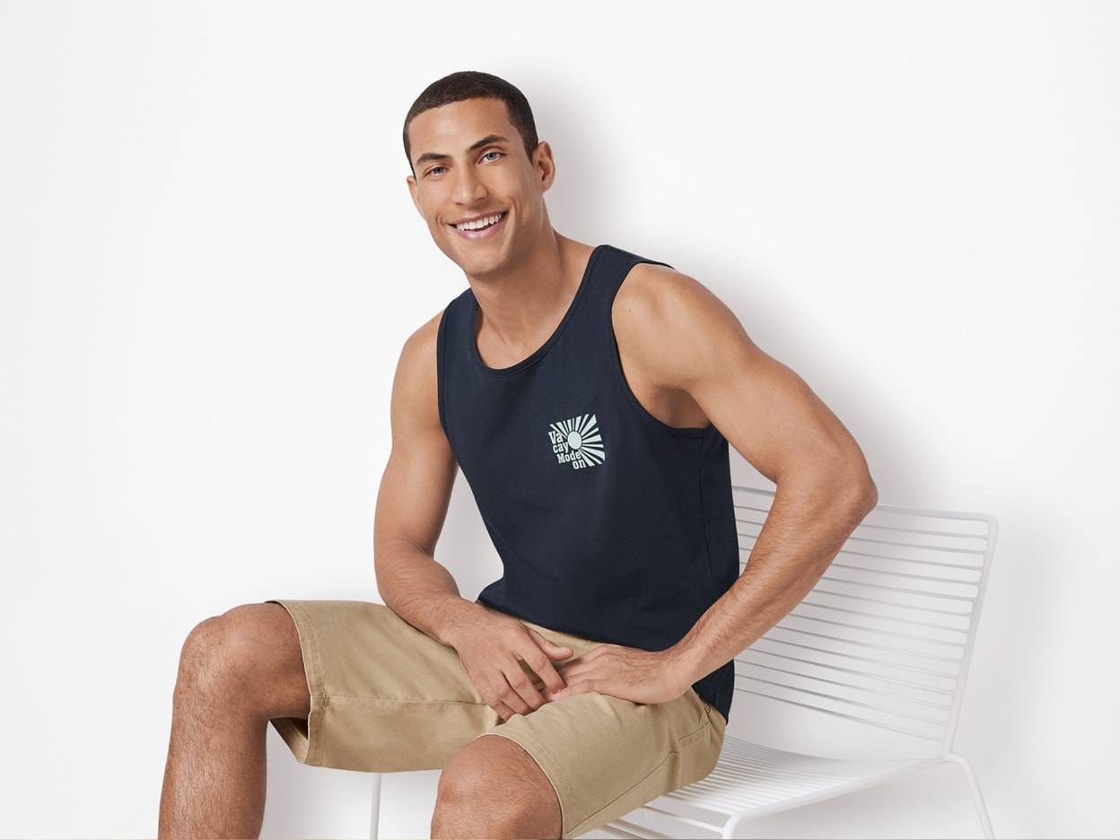 Ein lächelnder Mann trägt ein schwarzes Tanktop und eine beige Shorts.