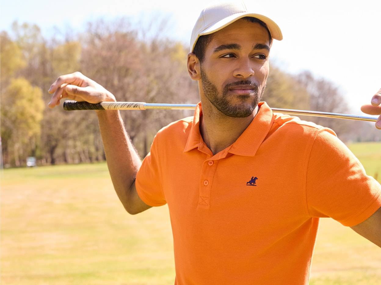 Ein Mann trägt ein orangefarbenes Polohemd und eine beigefarbene Kappe und trägt einen Golfschläger über der Schulter.