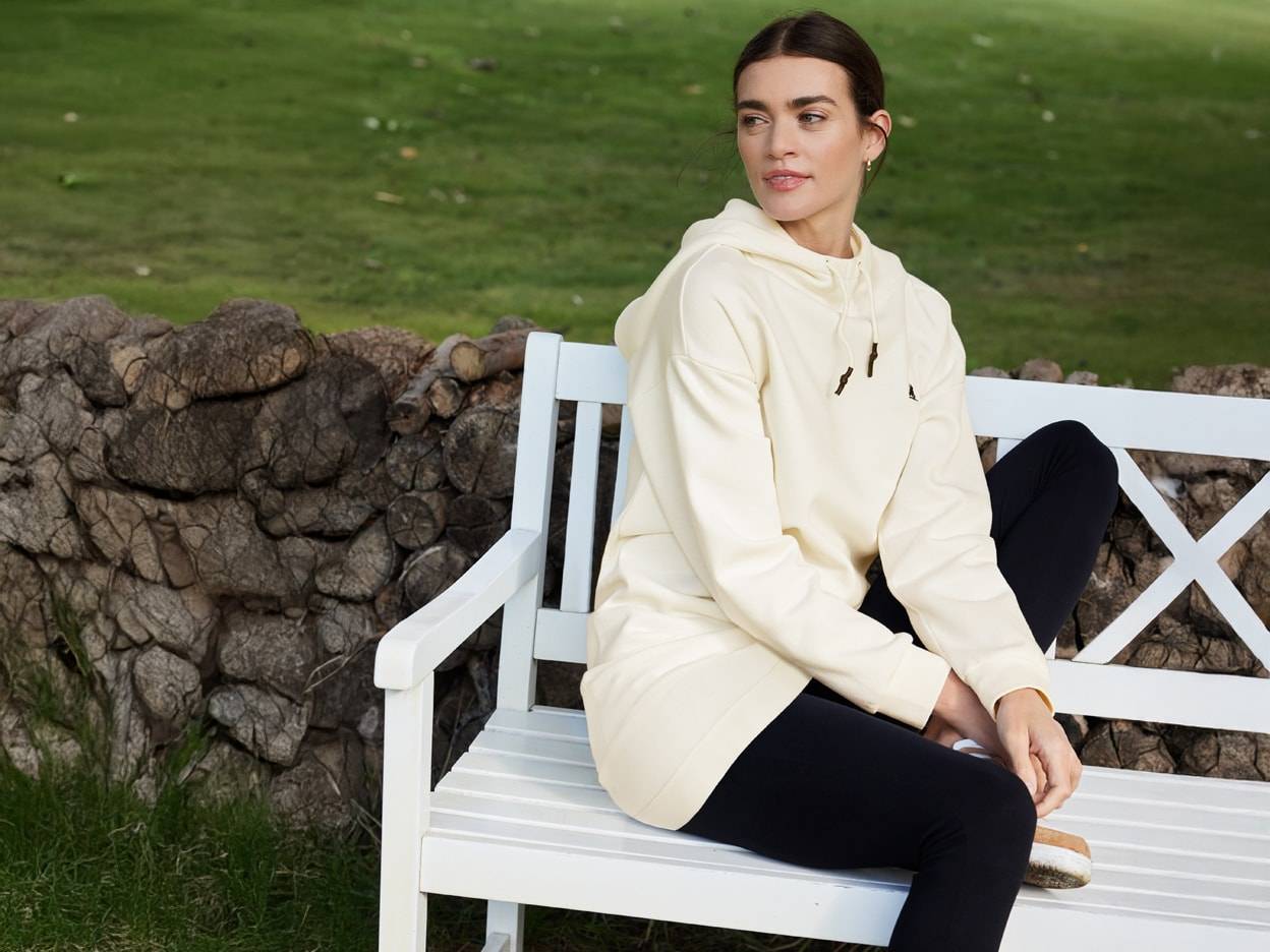 Ein Model trägt ein cremefarbenes Hoodie-Kleid und schwarze Leggings auf einer weißen Bank im Freien.