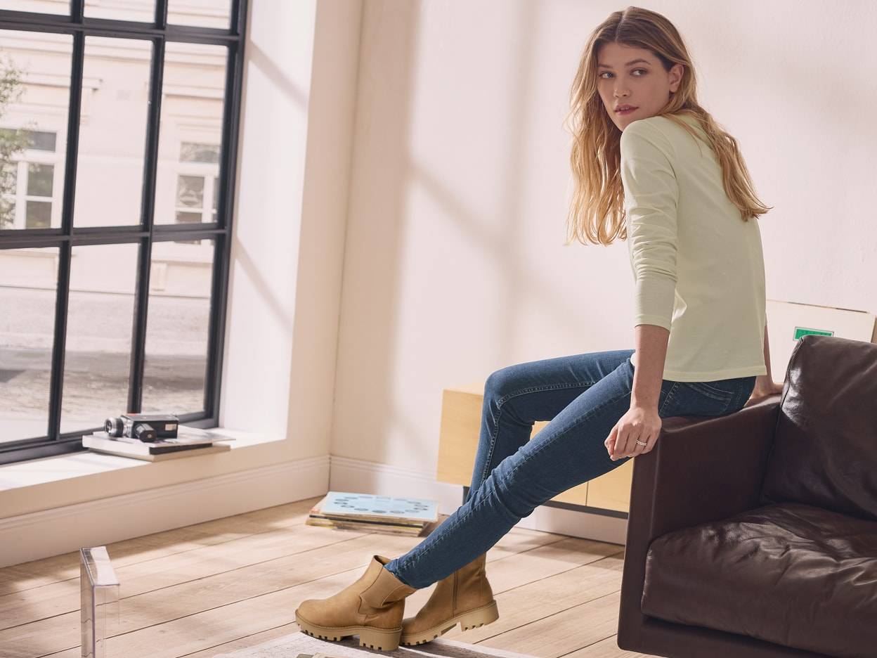 Eine Frau in Jeans und braunen Stiefeln sitzt auf einem Ledersofa, mit einem Fenster im Hintergrund.