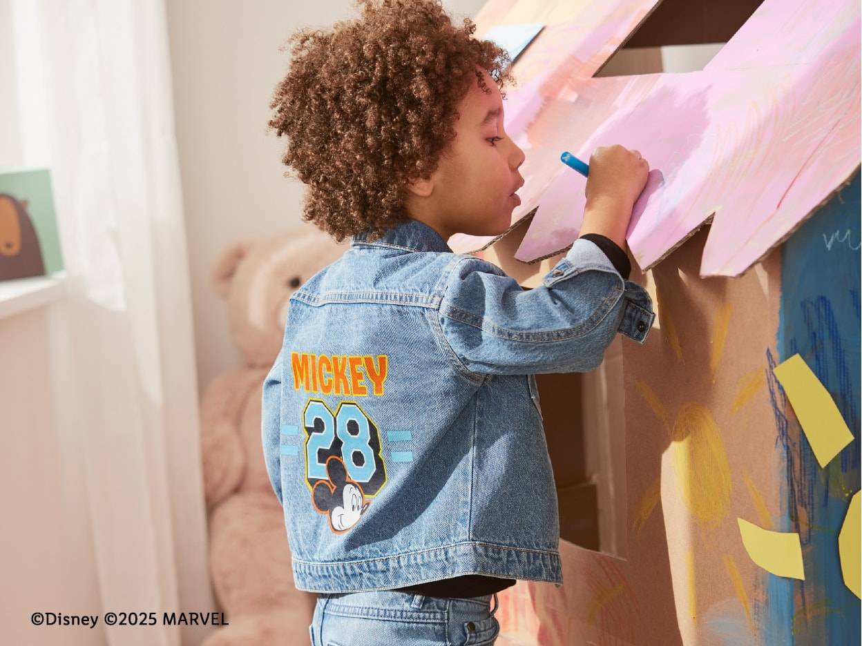 Un enfant porte une veste en jean Mickey Mouse et dessine sur une maison en carton.
