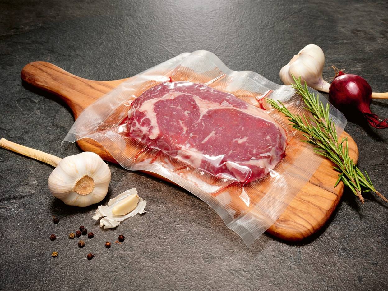 Ein Vakuumiertes Steak auf einem Holzbrett, mit Knoblauch, Rosmarin und schwarzen Pfefferkörnern.
